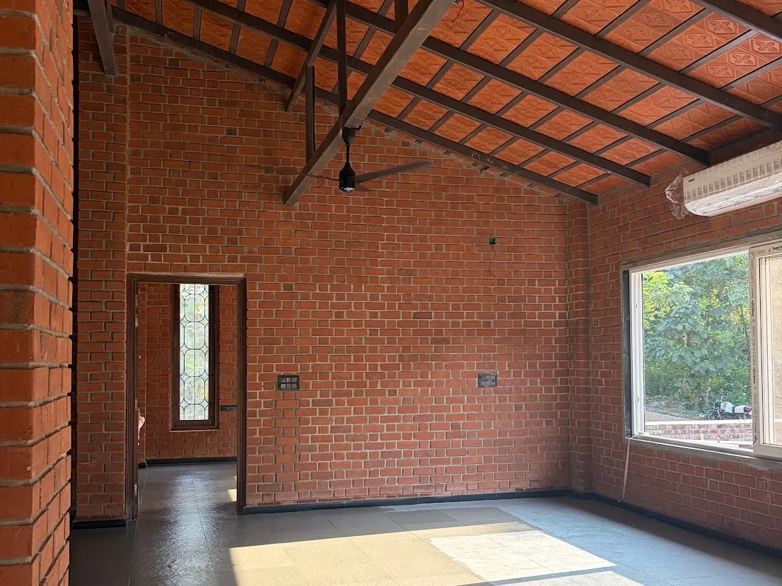 Inside the house under construction: exposed red-brick walls, a sloped tile roof on dark timber rafters, and a wide window onto the trees