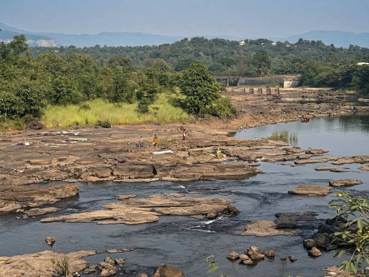A shallow river running over flat rocks in the Sahyadri hills, a few people and laundry drying on the rocks, forested hills behind